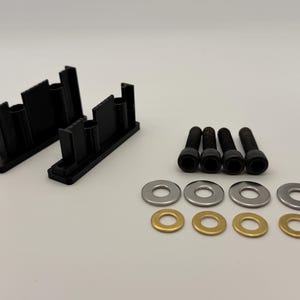 May include: Two black plastic mounting brackets with vertical supports, four black metal socket head cap screws, four silver-colored metal washers, and four brass-colored metal washers arranged in a set on a plain light background.