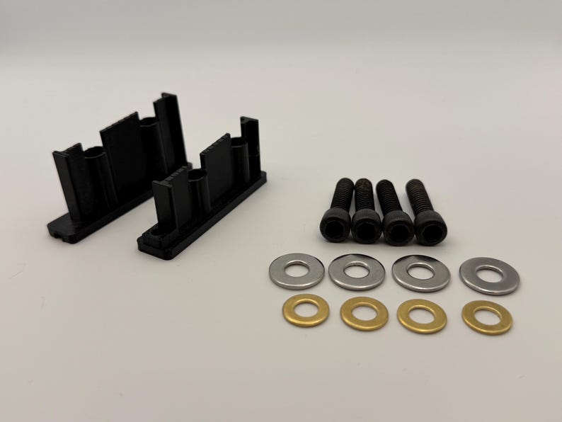 May include: Two black plastic mounting brackets with vertical supports, four black metal socket head cap screws, four silver-colored metal washers, and four brass-colored metal washers arranged in a set on a plain light background.