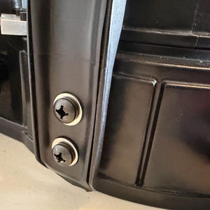 May include: A black plastic component featuring a vertical structural rib secured by two black Phillips head screws with silver washers. The surface has a smooth, molded finish with recessed rectangular paneling visible on the right side.
