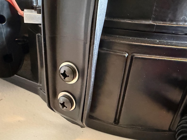 May include: A black plastic component featuring a vertical structural rib secured by two black Phillips head screws with silver washers. The surface has a smooth, molded finish with recessed rectangular paneling visible on the right side.