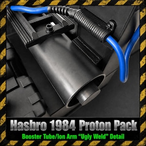 Puede incluir: Un componente de plástico negro de un Proton Pack Hasbro 1984 que presenta un tubo propulsor cilíndrico y un brazo iónico con un detalle de soldadura texturizado. Un cable azul brillante está conectado al conjunto, sobre un fondo negro.