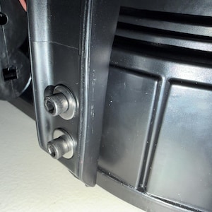 May include: A black subwoofer speaker component featuring a rigid plastic frame with a smooth finish. Two silver-toned metal hex bolts with washers are secured into the side of the frame, positioned vertically above one another on a flat surface.