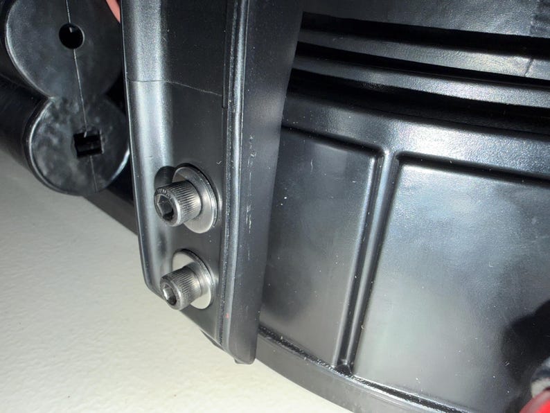 May include: A black subwoofer speaker component featuring a rigid plastic frame with a smooth finish. Two silver-toned metal hex bolts with washers are secured into the side of the frame, positioned vertically above one another on a flat surface.