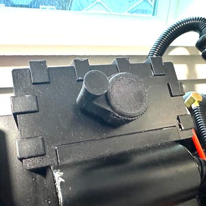 Puede incluir: Un dispositivo de plástico negro con una perilla redonda negra cubierta de malla y un mango cilíndrico negro. El dispositivo está conectado a una manguera negra.