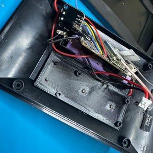 May include: An overhead view of the inside of a black electronic device, showing a circuit board with different coloured wires and components. A black rectangular component is secured with screws. Red and blue wires are visible.
