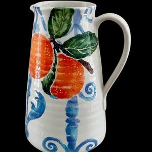 May include: White ceramic pitcher with a curved handle, decorated with orange fruit, green leaves, and blue floral patterns. The pitcher has a spout for easy pouring and is suitable for serving beverages.