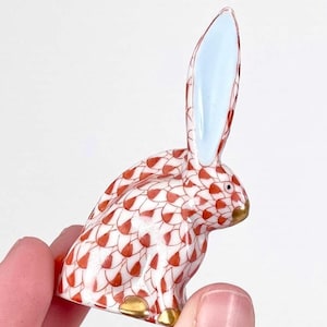 May include: A ceramic rabbit figurine with a white base and a red and white pattern. The rabbit's ears are light blue on the inside, and the feet and nose are gold. The figurine is held in a person's hand.