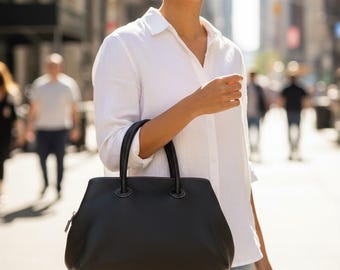 May include: A black handbag with a textured surface and two rounded handles. The bag has a zipper closure and a structured shape. The person in the image is wearing a white button-down shirt and blue jeans.