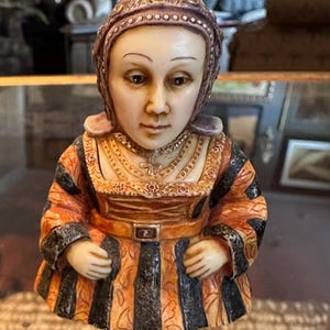 Peut inclure: Une figurine en céramique d'une femme portant une coiffe violette et dorée, un collier en or et une robe à rayures orange et noires.