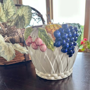 May include: White ceramic basket-shaped planter with a woven texture. The planter is decorated with colorful, raised relief grapes, cherries, and leaves. The grapes are a deep blue, the cherries are pink, and the leaves are green.