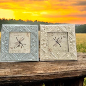 May include: Two decorative metal picture frames with a dragonfly design. The frames are in a light blue and white color. The frames are sitting on a wooden surface.