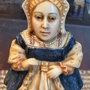 May include: A small, ivory-colored figurine of a woman wearing a blue and gold dress and a white head covering. The figurine is detailed with a pearl necklace and a white fabric tied around her waist.