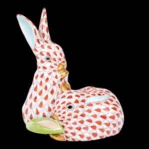 May include: A ceramic figurine of two rabbits. The rabbits are white with a red and gold pattern. The ears have a light blue interior. One rabbit is upright, and the other is lying down. A green leaf is between them.