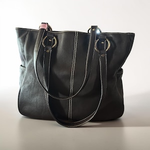 May include: A dark brown leather tote bag featuring two shoulder straps attached with large gold-tone metal rings. The bag has visible white contrast stitching along the center panel and straps, with side pockets on both the left and right sides.
