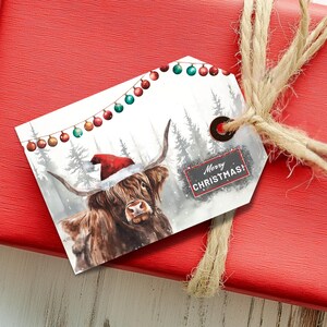 May include: A white gift tag with a watercolor illustration of a Highland cow wearing a Santa hat. The cow is standing in a snowy forest with a string of Christmas lights above. The tag says "Merry Christmas!" in a black frame.