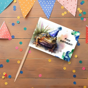 Könnte beinhalten: Eine Aquarell-Illustration eines Frosches mit einem blauen Partyhut auf einer weißen Karte mit dem Text "Happy Birthday!" Die Karte liegt auf einer Holzoberfläche mit bunten Konfetti.