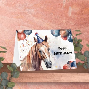 Może przedstawiać: Kartka urodzinowa z akwarelową ilustracją konia w czapeczce urodzinowej. Na kartce napis "Happy BIRTHDAY!" i dekoracja z czerwonych, niebieskich i szarych balon&oacute;w. Kartka leży na drewnianej p&oacute;łce.