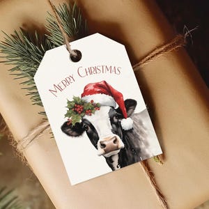 May include: A gift tag with a watercolor illustration of a cow wearing a Santa hat and holly. The words "Merry Christmas" are printed above the cow. The tag is tied to a wrapped gift with twine and a sprig of evergreen.
