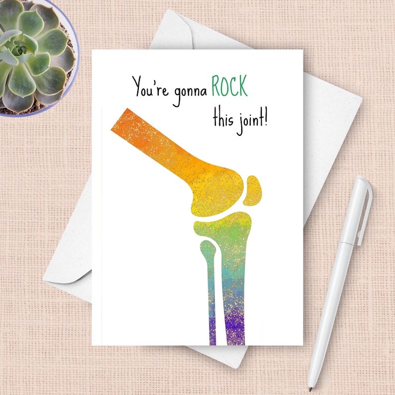 May include: A white greeting card with a colorful illustration of a knee joint. The text "You're gonna ROCK this joint!" is written in black and green. A white envelope and pen are also visible.