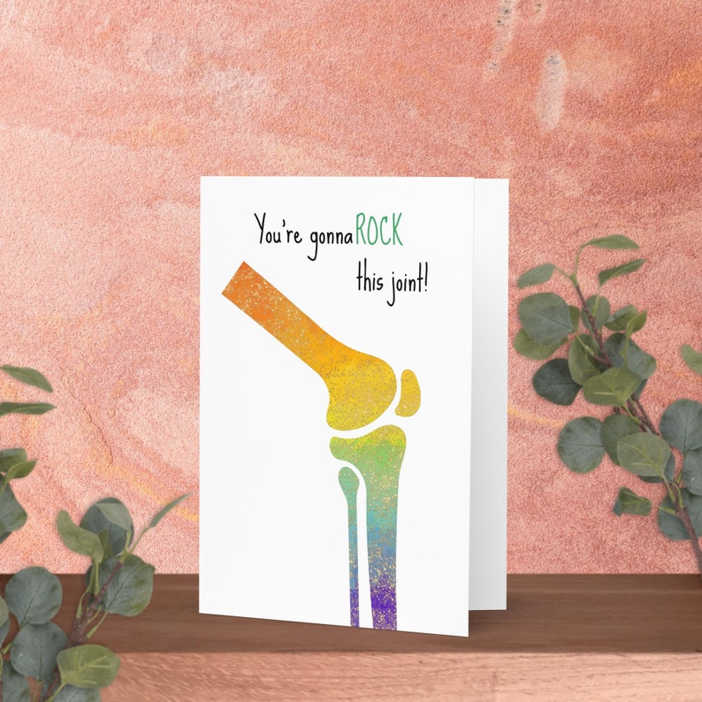 May include: A white greeting card with a colorful illustration of a knee joint. The text "You're gonna ROCK this joint!" is written in black and green. The card is displayed on a wooden surface.