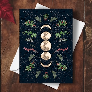 May include: Greeting card featuring a watercolor illustration of the moon phases against a dark blue starry background. The design includes botanical elements like green leaves, red berries, and sprigs of evergreen. The card is on a white envelope, set on a wooden surface.