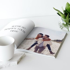 May include: An open photo book with a picture of a person giving another person a piggyback ride on a beach. The book is next to a white mug on a white marble coaster. The text "WE'RE SO HAPPY YOU'RE HERE" is printed on the left page.