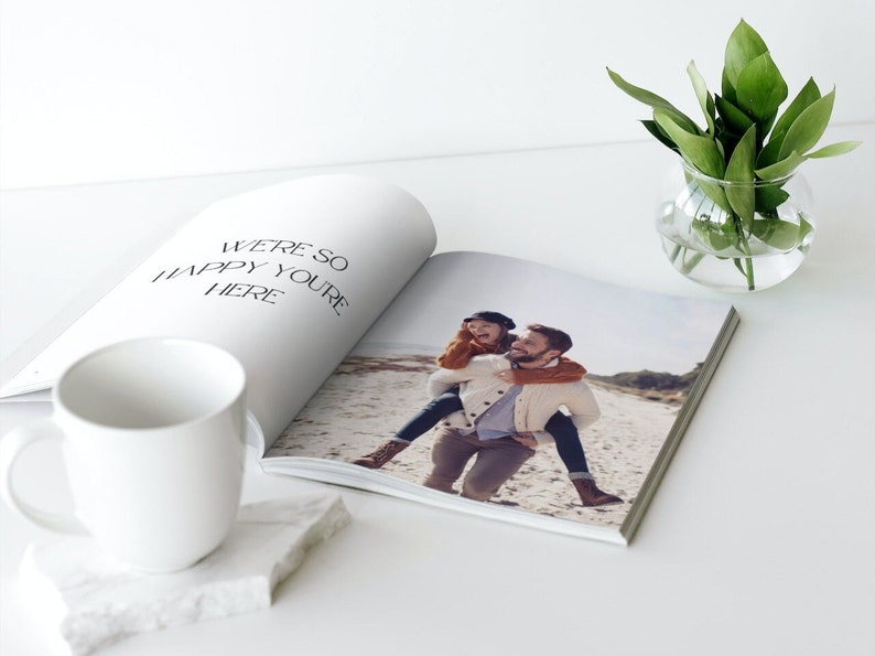 May include: An open photo book with a picture of a person giving another person a piggyback ride on a beach. The book is next to a white mug on a white marble coaster. The text "WE'RE SO HAPPY YOU'RE HERE" is printed on the left page.