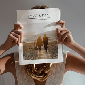May include: A person holding a wedding photo album with the names "EMMA & DAN" and the date "12 JANUARY 2024 | WHISTLER, CANADA". The album shows a couple walking on a wooden walkway towards the sea. A gold chain necklace is visible.