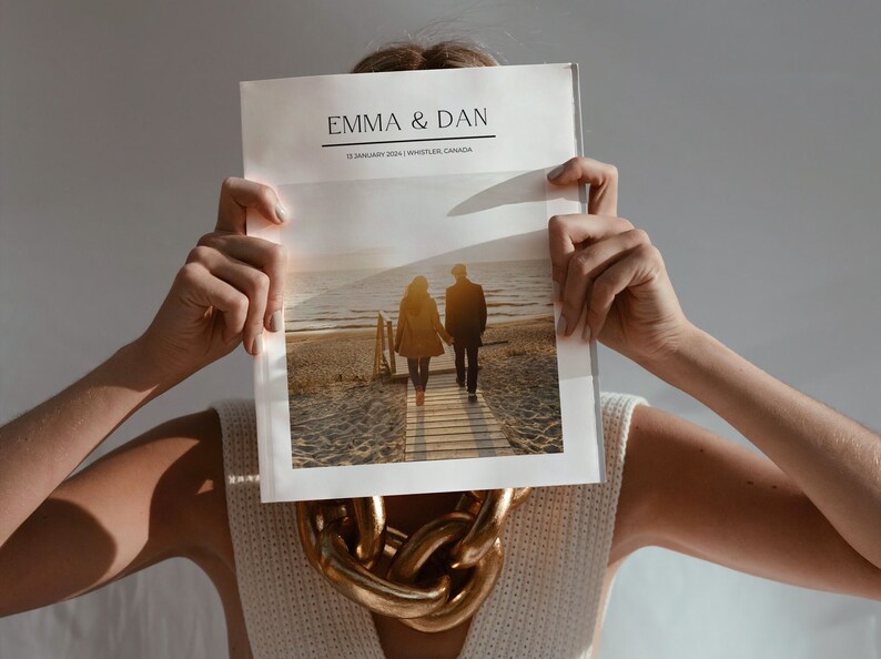 May include: A person holding a wedding photo album with the names "EMMA & DAN" and the date "12 JANUARY 2024 | WHISTLER, CANADA". The album shows a couple walking on a wooden walkway towards the sea. A gold chain necklace is visible.