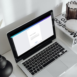 May include: A silver laptop displaying the text "HOW WE MET" on its screen sits on a white desk. A black keyboard is visible. A black vase with a dried plant and a candle holder with a candle are also on the desk.