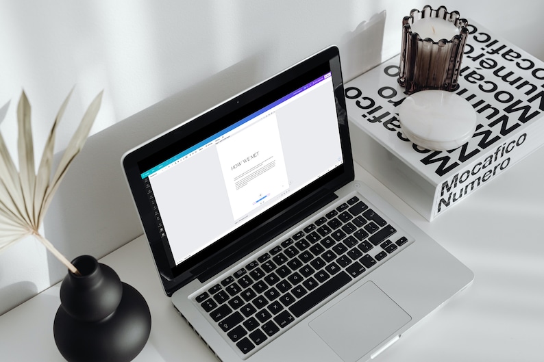 May include: A silver laptop displaying the text "HOW WE MET" on its screen sits on a white desk. A black keyboard is visible. A black vase with a dried plant and a candle holder with a candle are also on the desk.