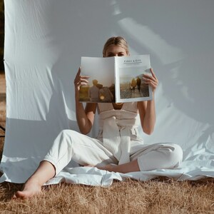 May include: A person seated, holding open a magazine with the text "EMMA & DAN" visible. The magazine features photographs of people. The person is wearing a cream-coloured outfit. The background is a white sheet and dry grass.