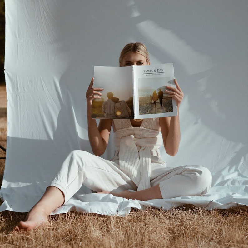 May include: A person seated, holding open a magazine with the text "EMMA & DAN" visible. The magazine features photographs of people. The person is wearing a cream-coloured outfit. The background is a white sheet and dry grass.