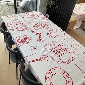 May include: A white rectangular tablecloth featuring a red Christmas pattern. The design includes illustrations of gifts, candy canes, a snowman, a sweater, and a wreath. The tablecloth is draped over a rectangular table with black chairs.