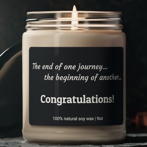 May include: A beige soy candle with a black label that reads "The end of one journey... the beginning of another... Congratulations! 100% natural soy wax | 9oz". The candle is lit and the flame is visible.