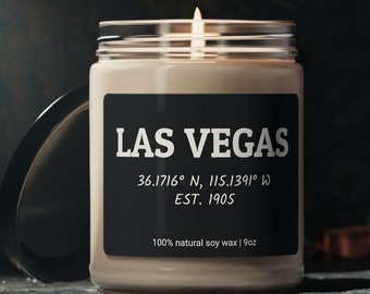 May include: A beige candle with a black label that reads "LAS VEGAS 36.1716° N, 115.1391° W EST. 1905 100% natural soy wax | 9oz". The candle is lit and the flame is visible.