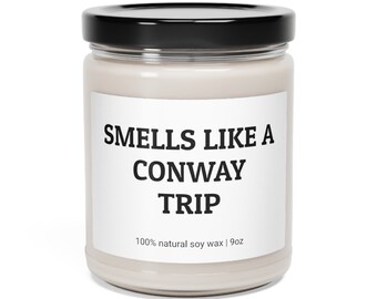 May include: A white candle jar with a black lid. The label reads "Smells Like A Conway Trip" and "100% natural soy wax | 9oz".