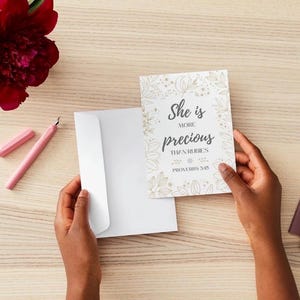 May include: A greeting card with the text "She is more precious than rubies" and floral accents is held near a white envelope. A pink pen and a red flower are also on the wooden surface.