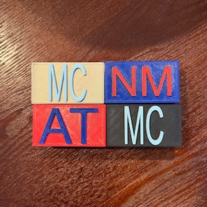 Può includere: Quattro blocchi quadrati con lettere che compongono "MCAT NM MC" in lettere blu, rosse e bianche su uno sfondo beige, blu, rosso e nero.