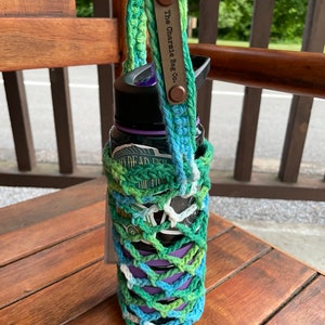 May include: A green, blue, and white crocheted bottle holder with a brown leather strap that says "The Charmed Bag Co."