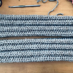 May include: Two grey, hand-crocheted items, possibly boot cuffs or leg warmers, are laid out on a wooden surface. The items are made with a thick yarn, showing a ribbed texture. A crochet hook and yarn are also visible.