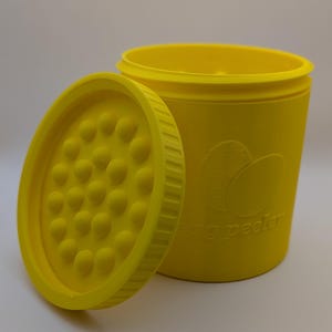May include: Bright yellow egg peeler with a matching lid. The lid is off to the side, revealing a textured surface. The container has an embossed design of two eggs and the words "egg peeler".