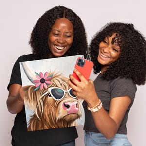 May include: Two people are holding up a painting of a Highland cow wearing sunglasses and a pink flower. The cow is looking to the right. The people are smiling and looking at the painting.