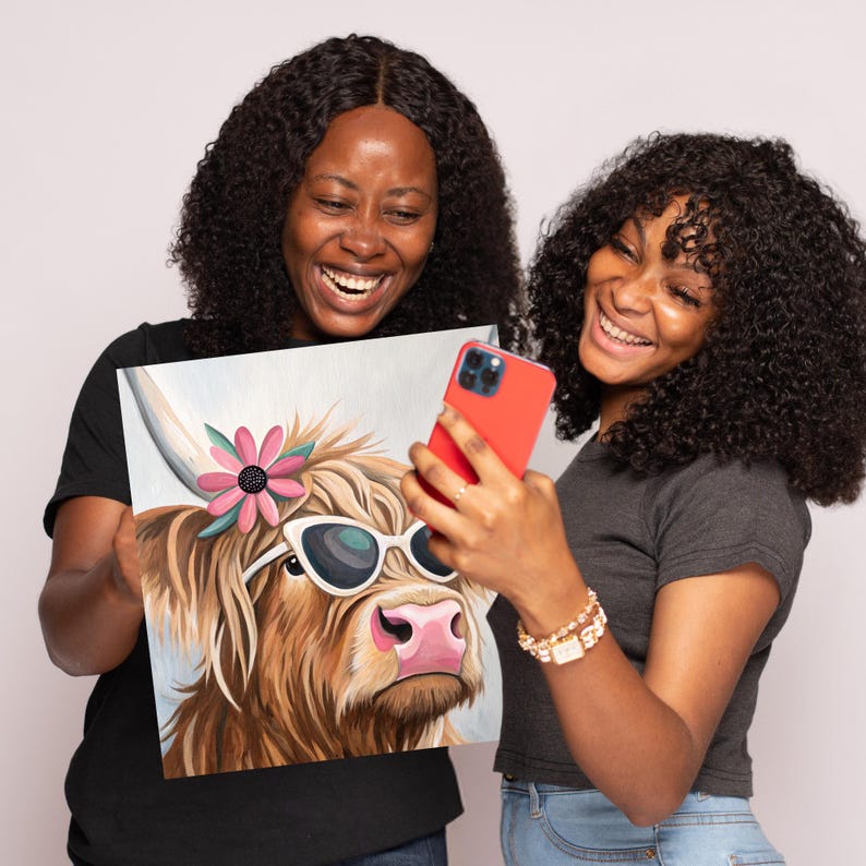 May include: Two people are holding up a painting of a Highland cow wearing sunglasses and a pink flower. The cow is looking to the right. The people are smiling and looking at the painting.