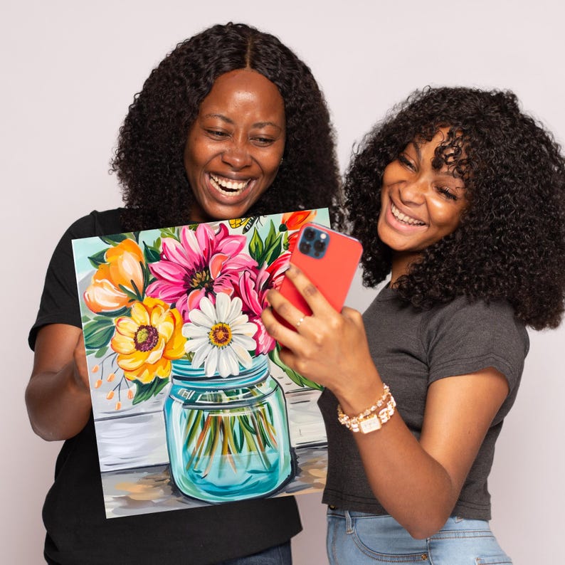 May include: Two people are smiling and looking at a phone. One person is holding a painting of a mason jar filled with colorful flowers. The painting has a white background with a wooden texture.