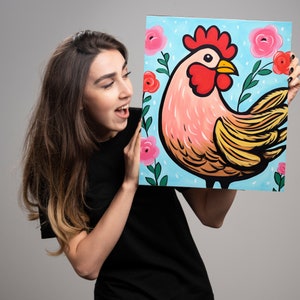 May include: A woman is holding a painting of a rooster with a pink body and yellow feathers. The rooster is on a blue background with pink flowers.