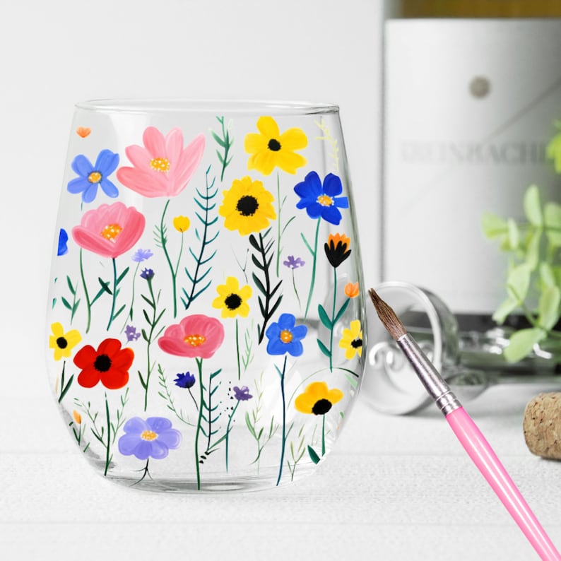 May include: A clear glass wine glass with a hand-painted design of colorful flowers. The flowers are pink, red, yellow, blue, and green. The glass is sitting on a white surface.