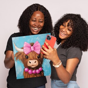 May include: Two people are holding up a painting of a brown and white Highland cow with a pink bow and pink beads. The cow is looking at the camera. The people are smiling and looking at a phone.