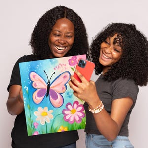 May include: Two people are smiling and holding a canvas painting of a pink butterfly with purple accents, surrounded by flowers in shades of pink, white, and yellow. The background is a light blue and green gradient.