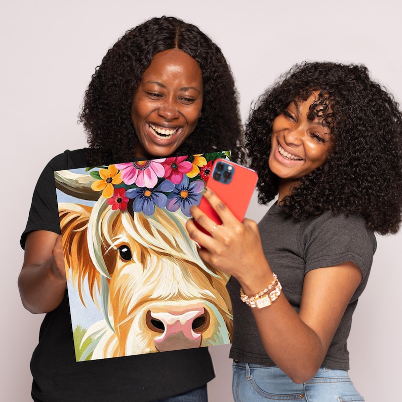 May include: Two people are holding up a painting of a Highland cow with a flower crown. The cow is white and brown with a pink nose. The painting is on a canvas and has a white background.